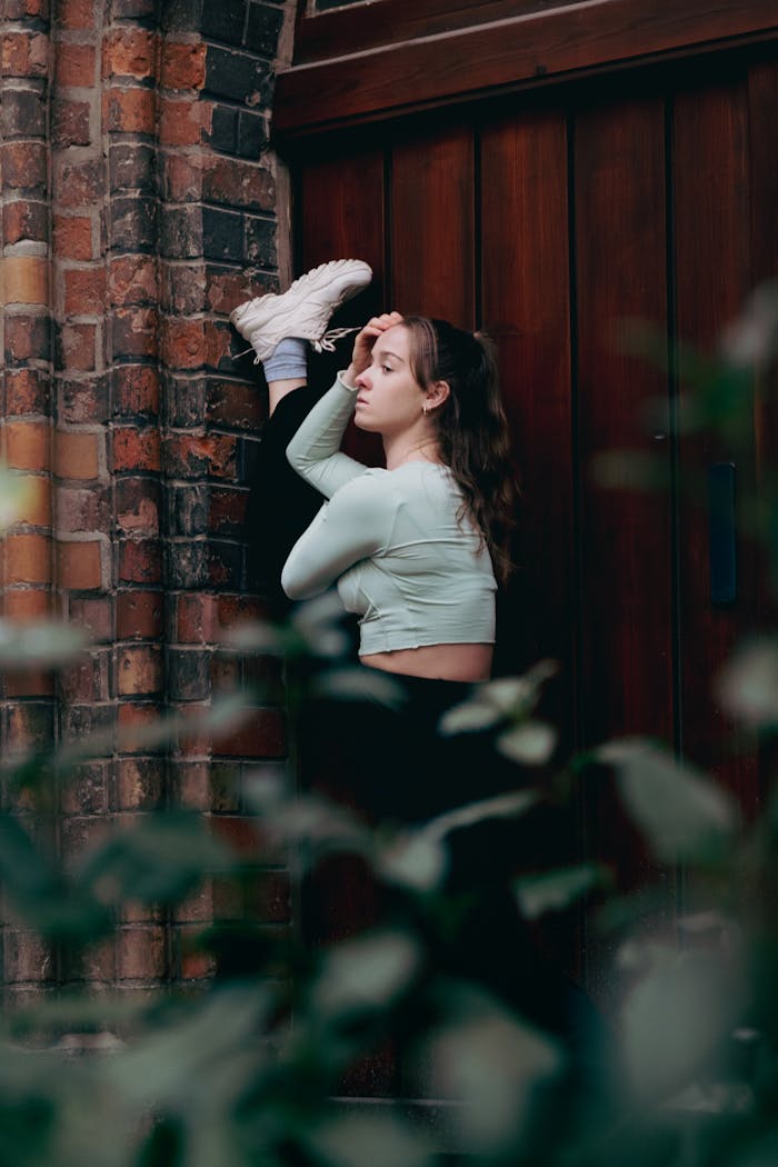 A young woman performs a high-leg stretch against a brick wall, showcasing flexibility and fitness.