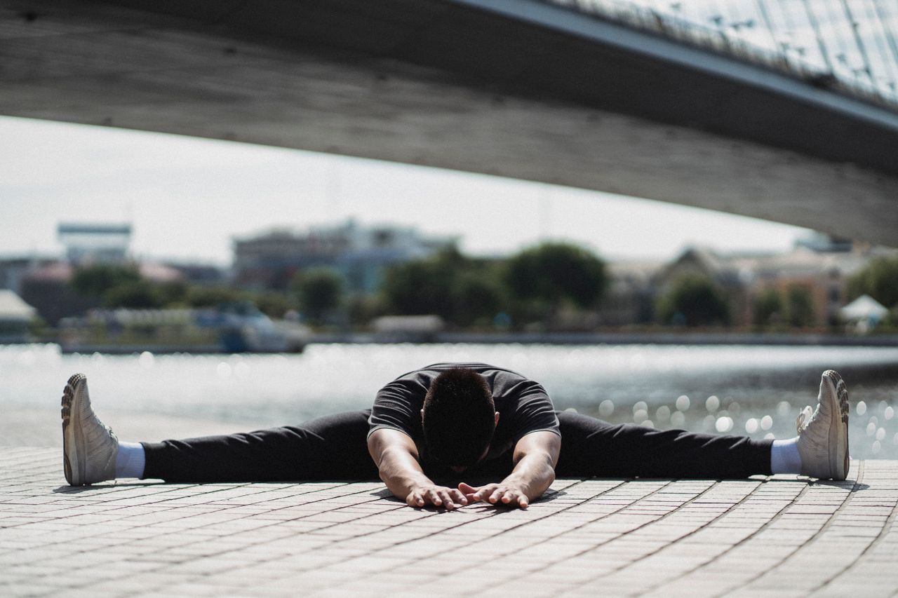 Ground level of anonymous flexible male sitting in Wide Legged Forward Bend pose while practicing yoga near bridge in town
